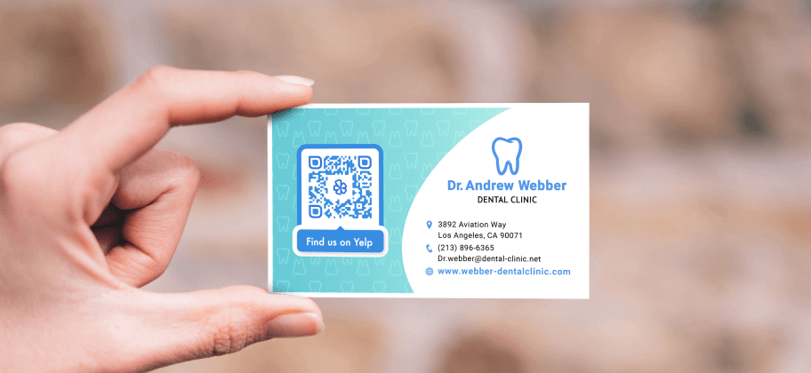 A hand holding up a business card from a dental clinic, including contact information and a QR Code for Yelp reviews.